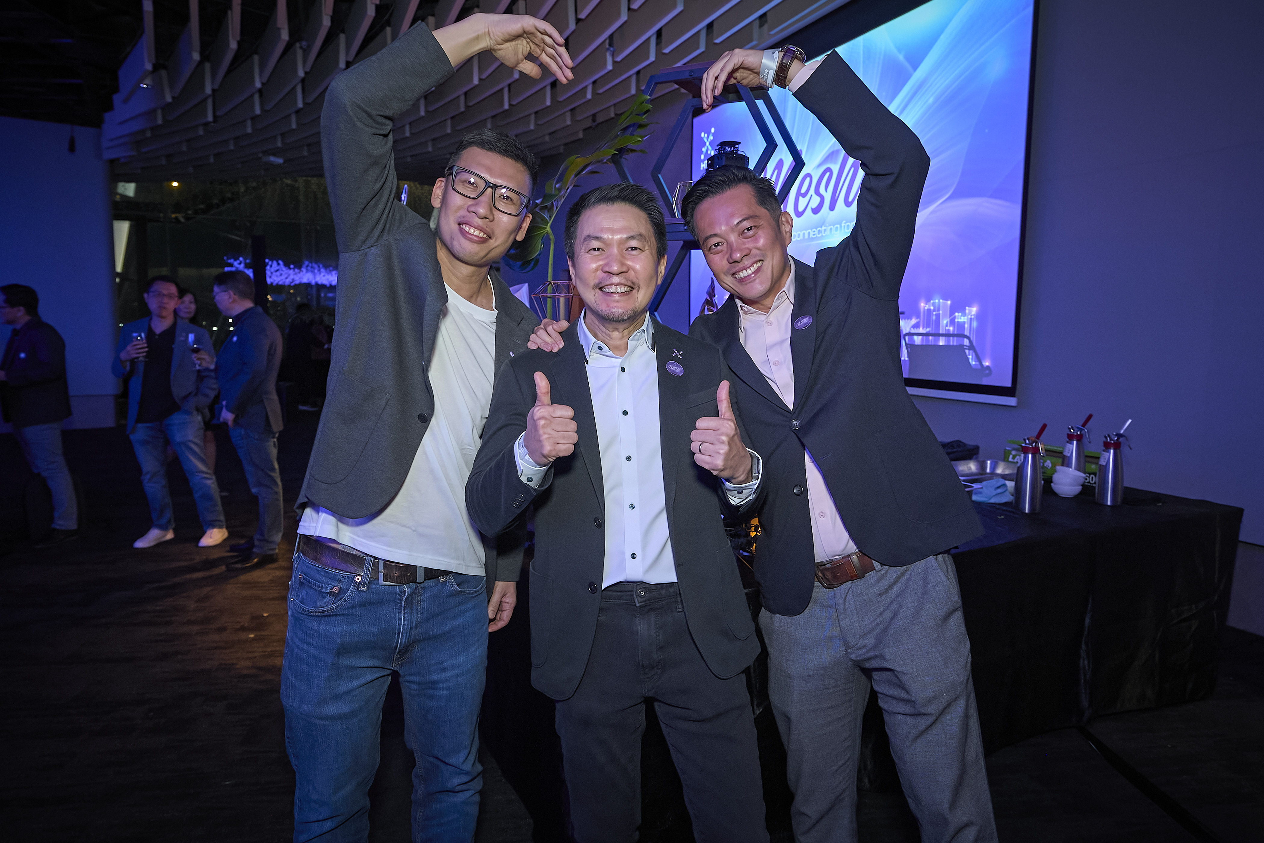Three men pose cheerfully, with the middle giving thumbs up and the other two forming heart shapes.