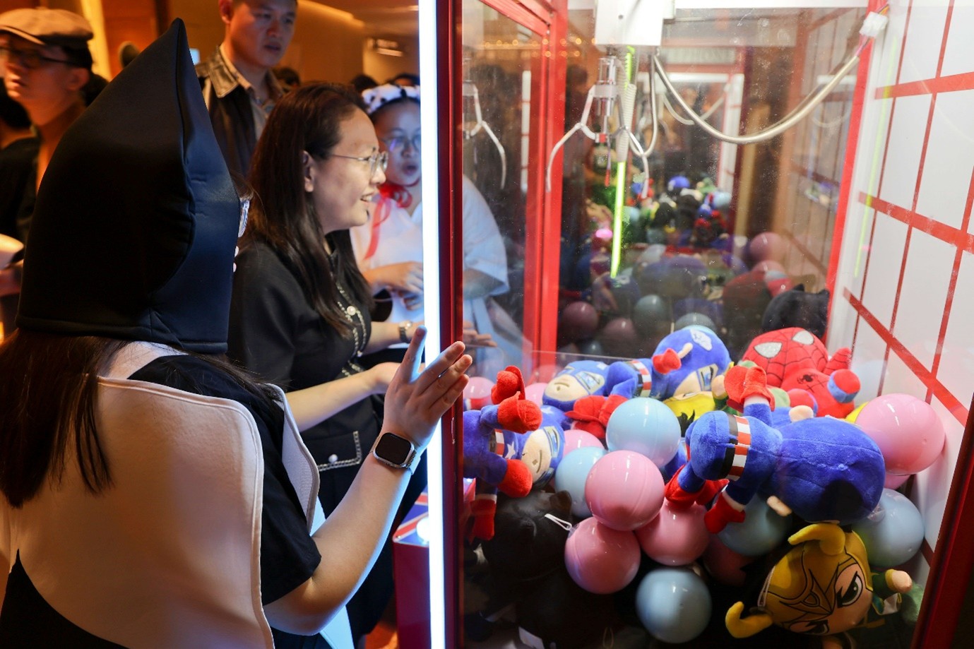 A group of people looking at a claw machine with superhero plush toys in them.