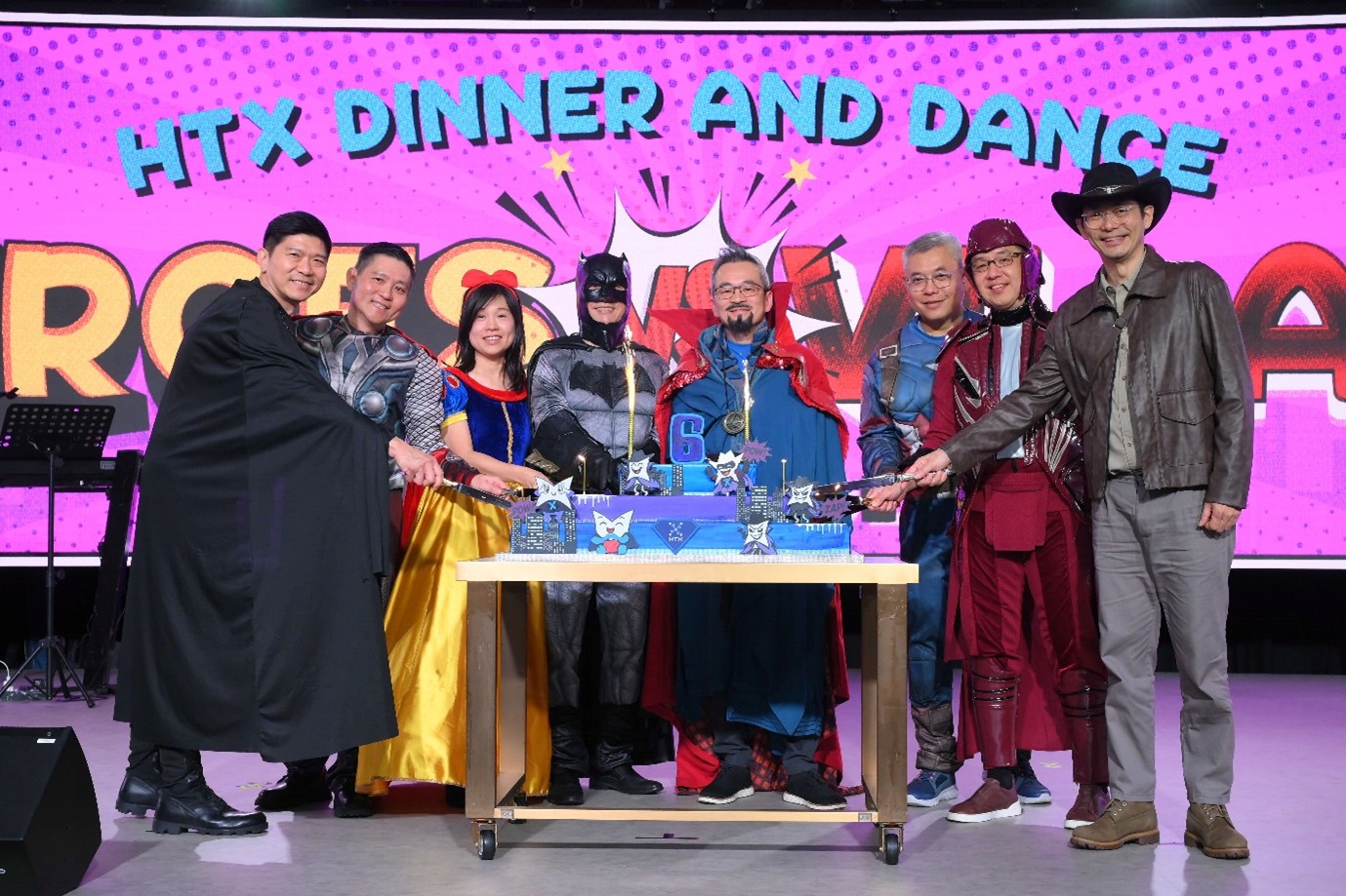 A group of people in superhero clothing stand around a large cake on a table, which has the number '6' and candles on it.