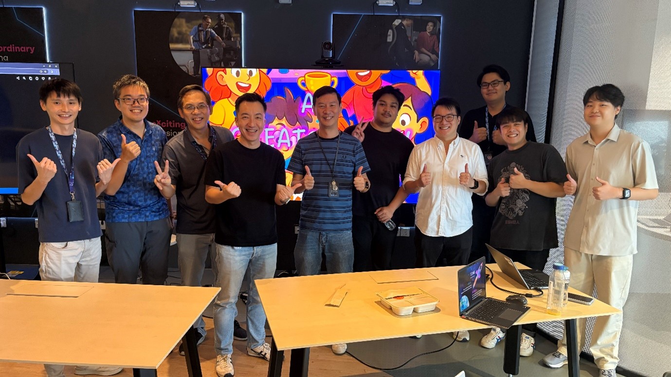 A group of people standing in front of a colourful TV screen and posing with their thumbs up.