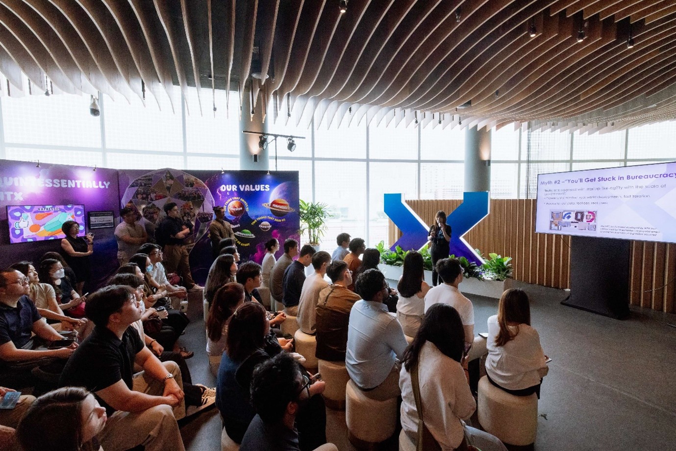 A standing woman holding a microphone in front of a seated audience.