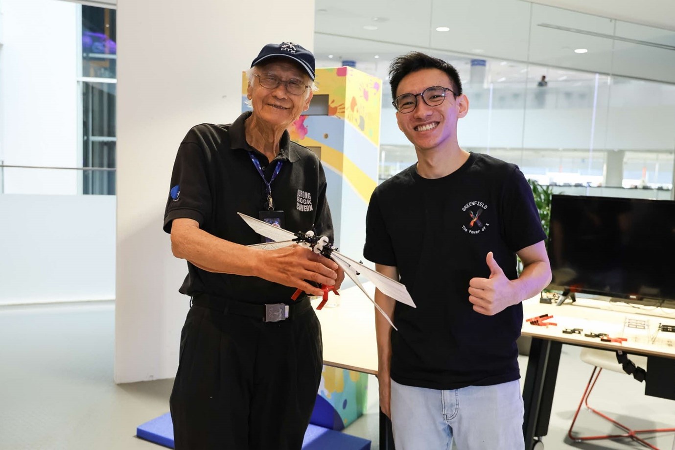 Two men smiling for the camera as one holds a robot with wings.