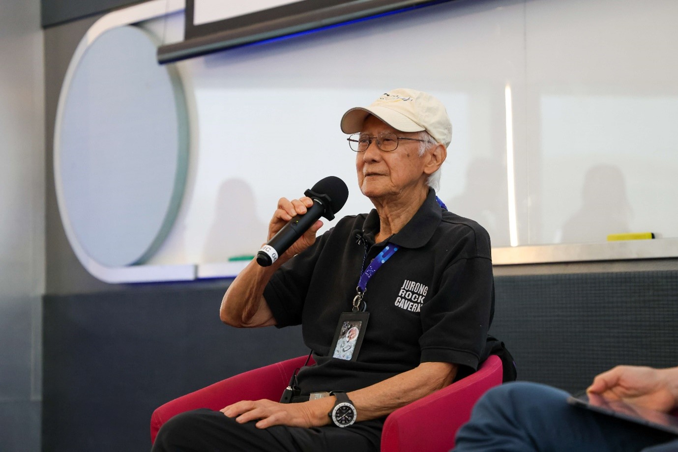 A person in a cap seated and holding a microphone.