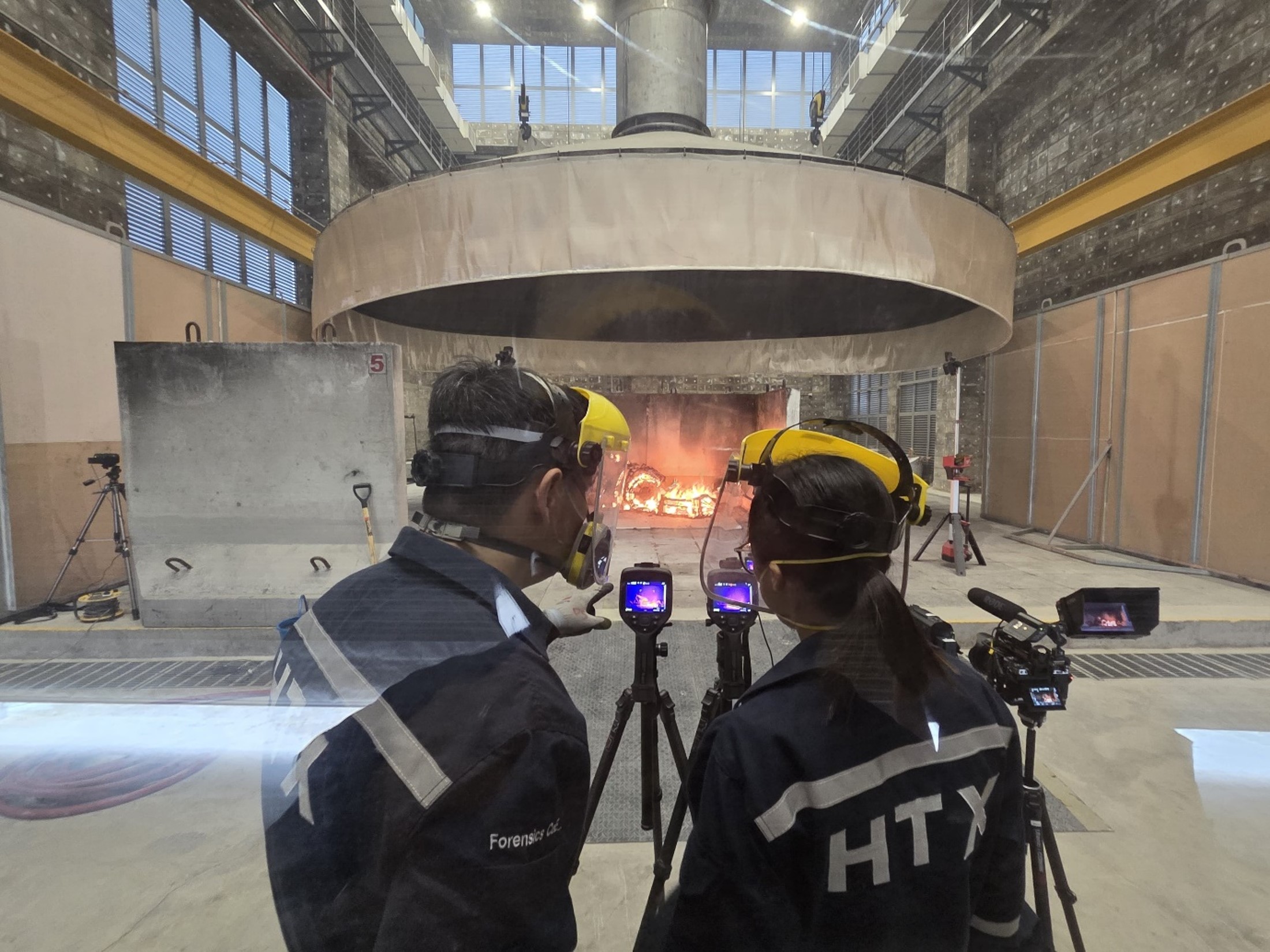 Two people in overalls and protective facial gear stand looking into cameras pointed at an object in the distance burning in a furnace set-up.