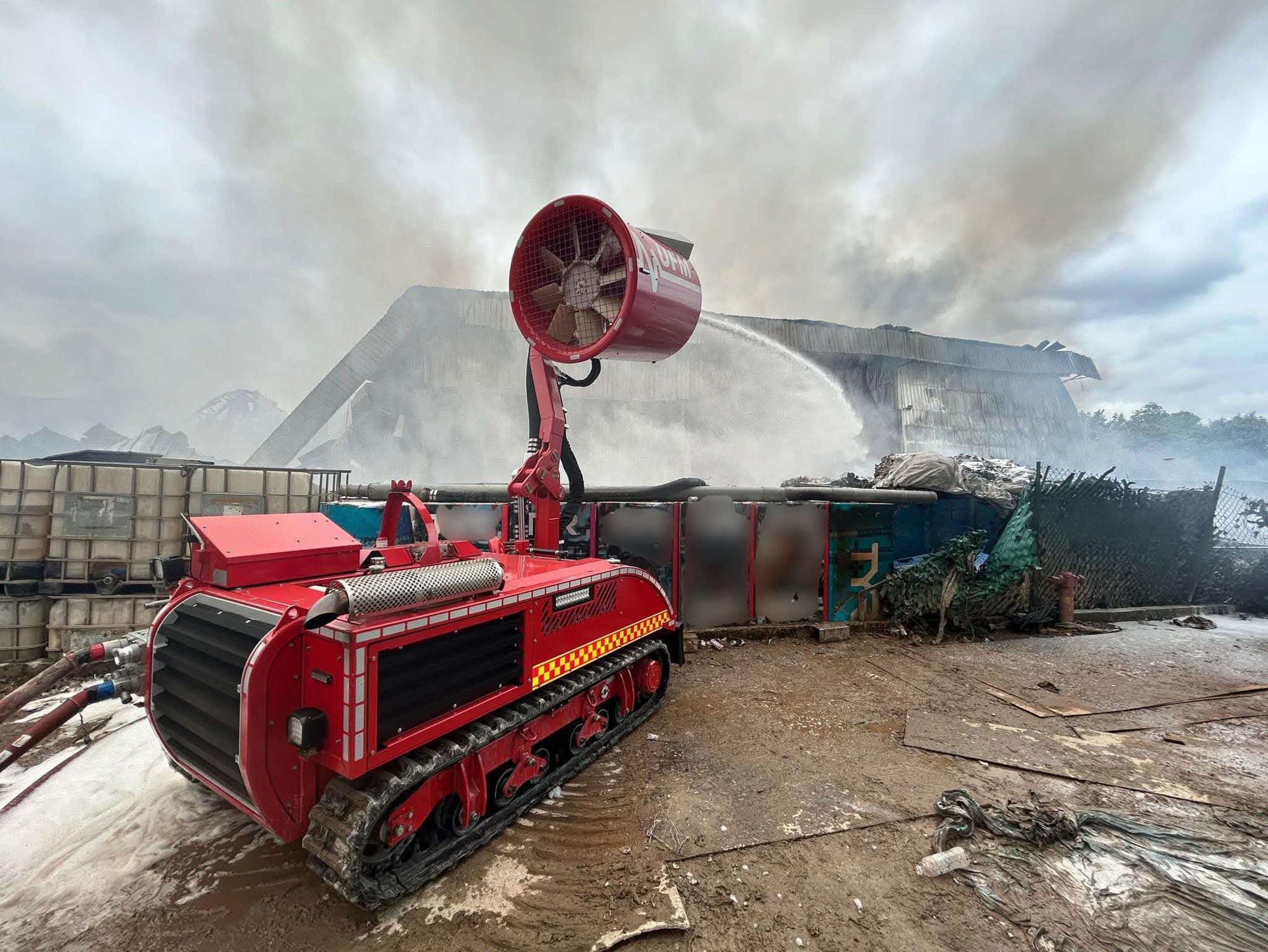 A large machine with rollers and a large fan spraying water at a burning building.