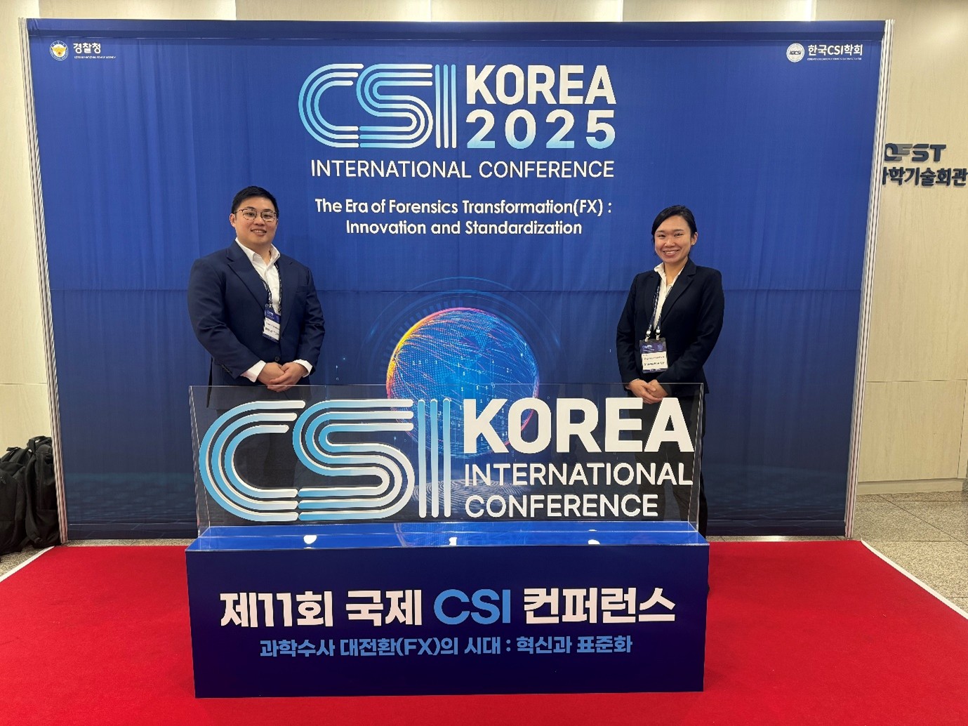 A man and a woman in business attire stand on a red carpet in front of a blue “CSI Korea 2025 International Conference” backdrop.