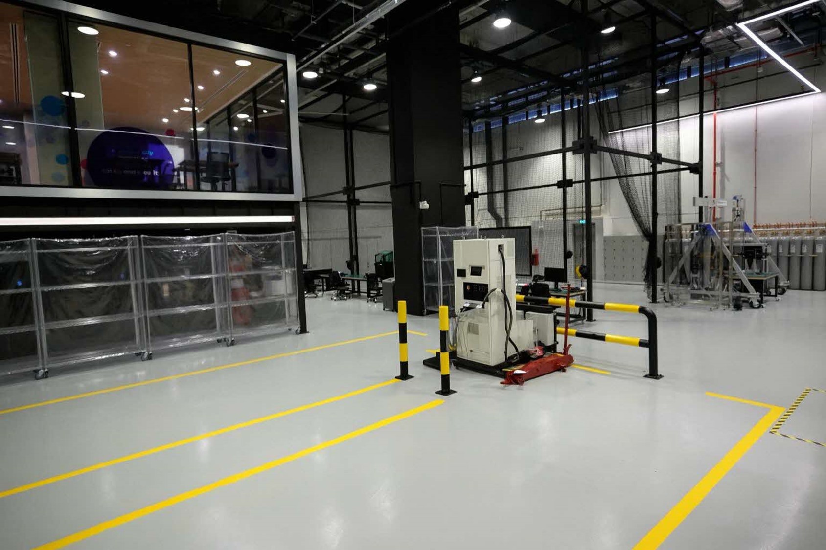 A large room with yellow lines marking out parking lots, yellow and black poles for vehicles, an electric vehicle charger in the centre, a large net-like structure in the back and a mezzanine room.