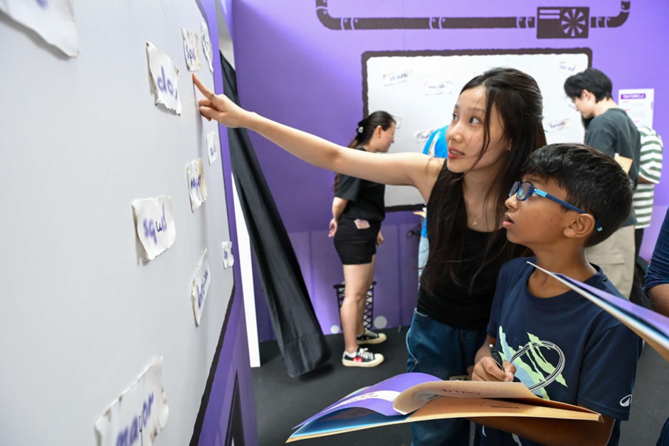 A woman points to words written on small pieces of paper stuck to a wall, while a young boy with glasses looks on holding a pen and notebook.