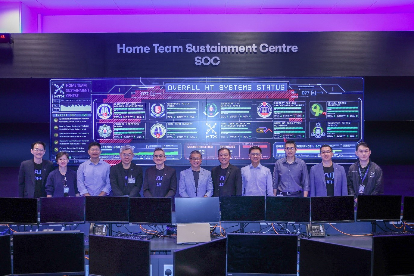 Group photo at the Home Team Sustainment Centre with system status screens in the background.