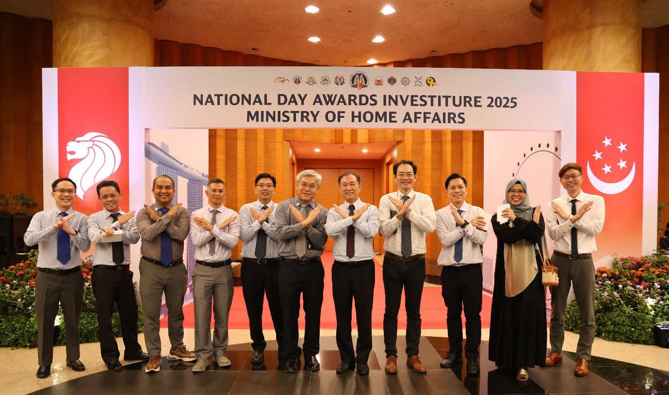 A group of people in formal wear pose with their arms crossed in an X shape while standing in front of a red-and-white arch which reads "National Day Awards Investiture 2025 Ministry of Home Affairs".
