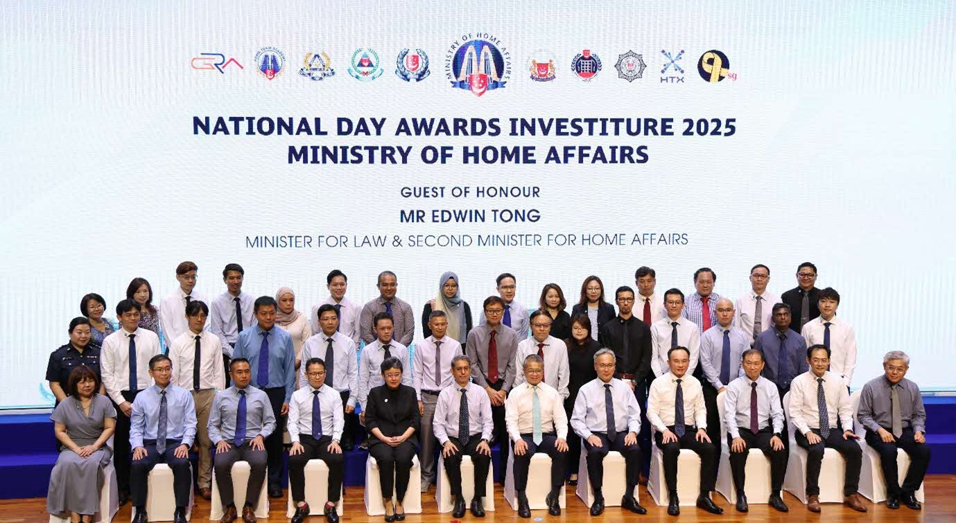 A formal group photo of a large group of people on stage in front of a backdrop which reads "National Day Awards Investiture 2025 Ministry of Home Affairs".
