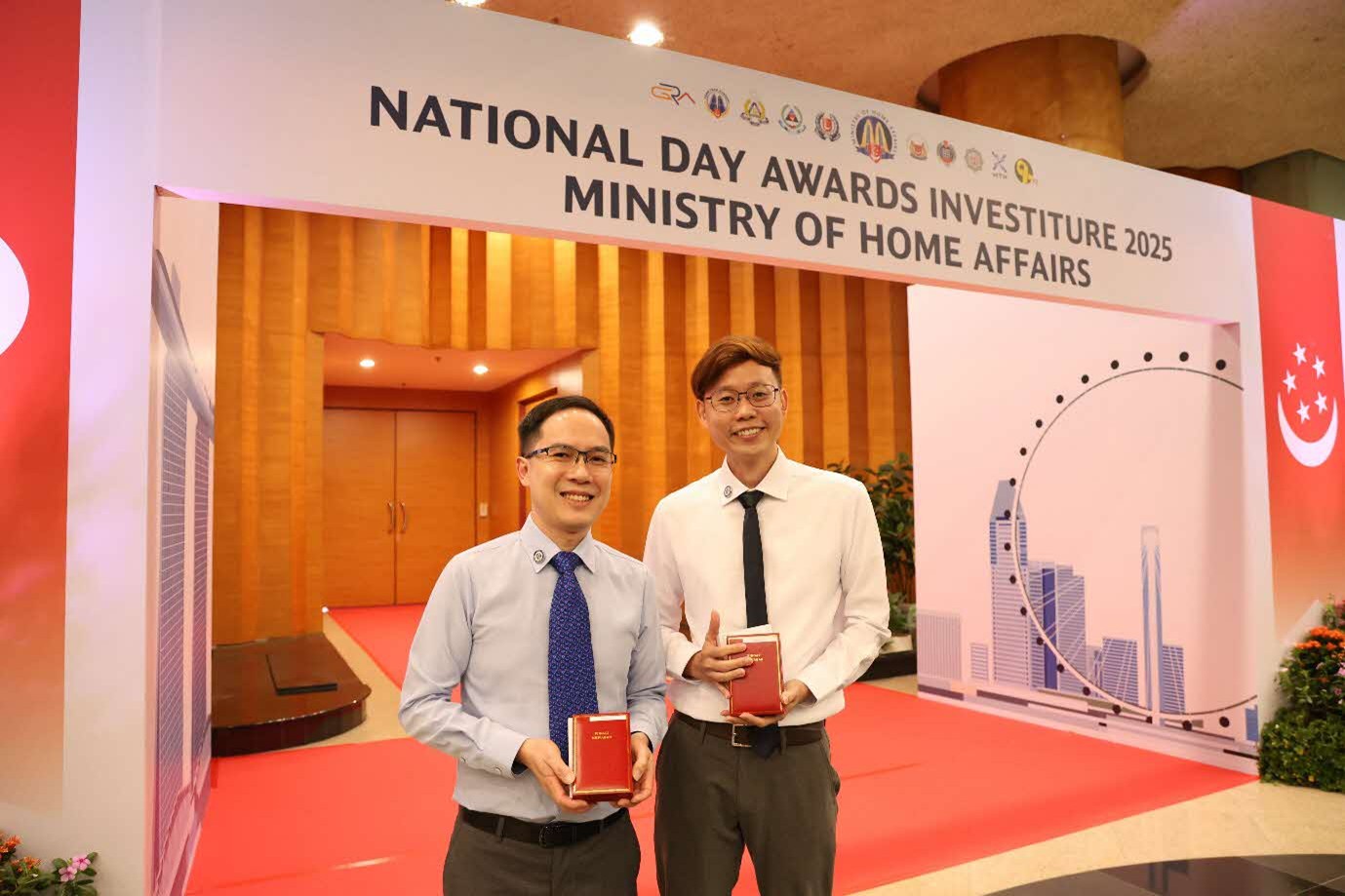 Two men in formal wear hold boxes and smile while standing in front of a large red-and-white arch that reads "National Day Awards Investiture 2025 Ministry of Home Affairs".
