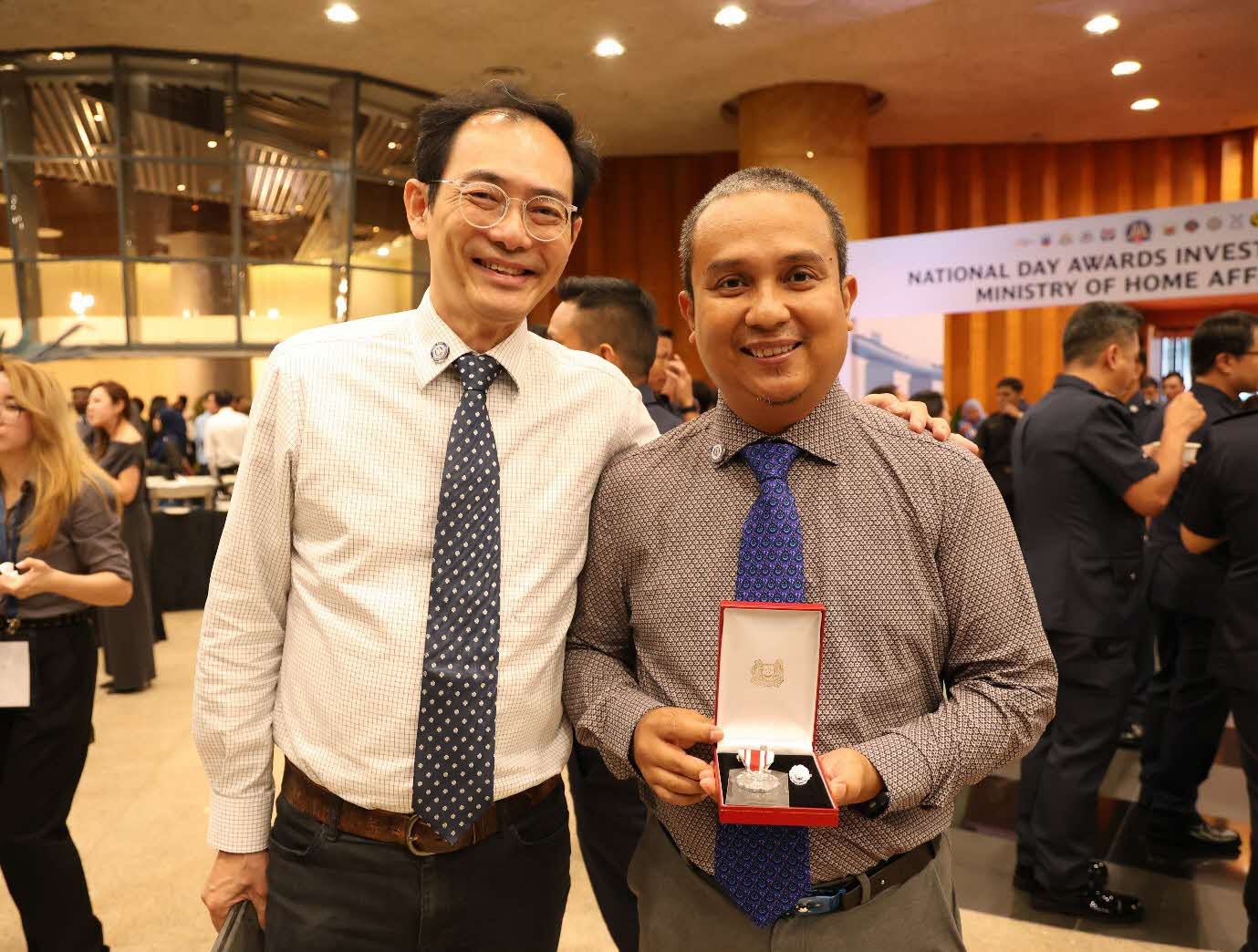 Two men in formal wear smile at the camera, with one holding an opened box with a medal in it.