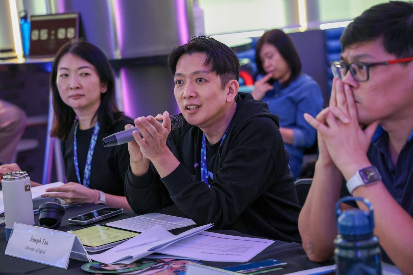 A judge speaking into a microphone during Prompt Royale 2025, with other judges seated beside him.