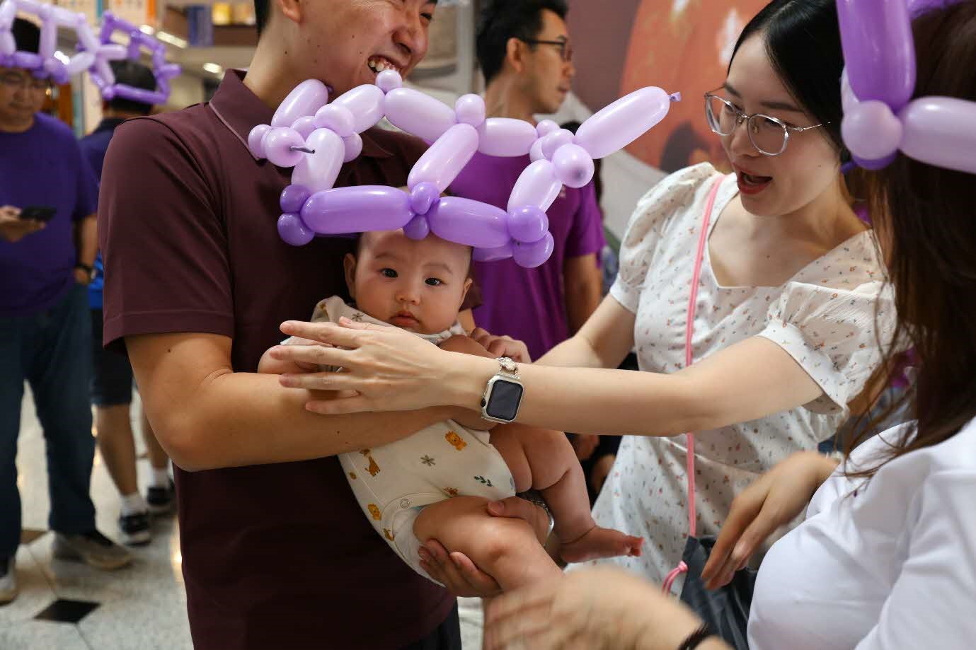 A baby being held by two people with a balloon hat on its head.