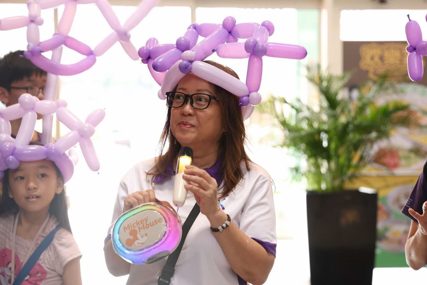 A woman wearing a balloon hat and holding a microphone and portable speaker.