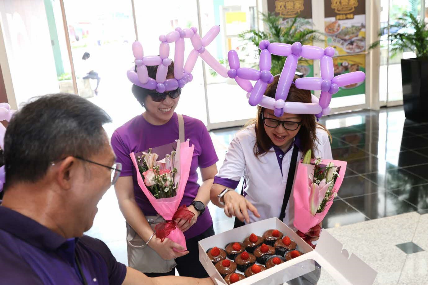 Two females holding flower bouquets and wearing balloon hats while looking at a box of cupcakes.