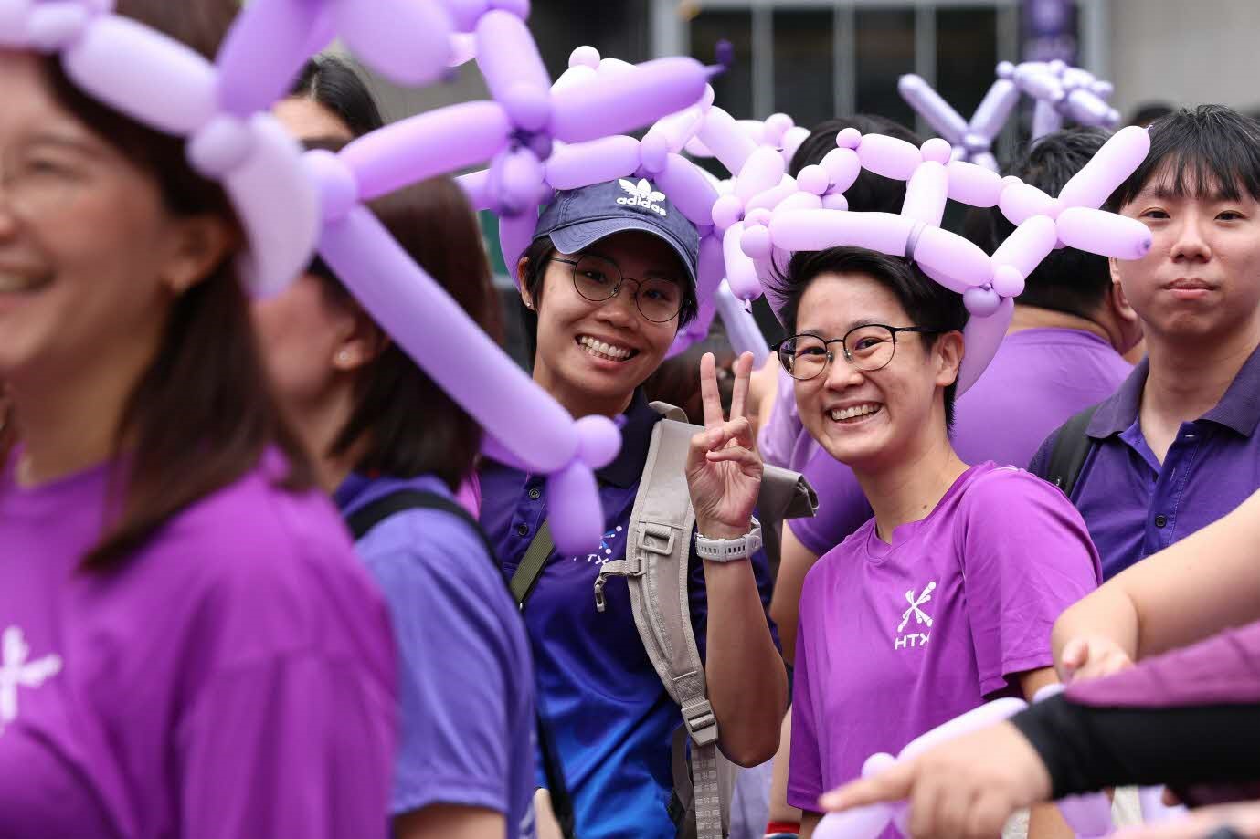 Two people smile amidst a group of people wearing purple shirts and balloon hats.