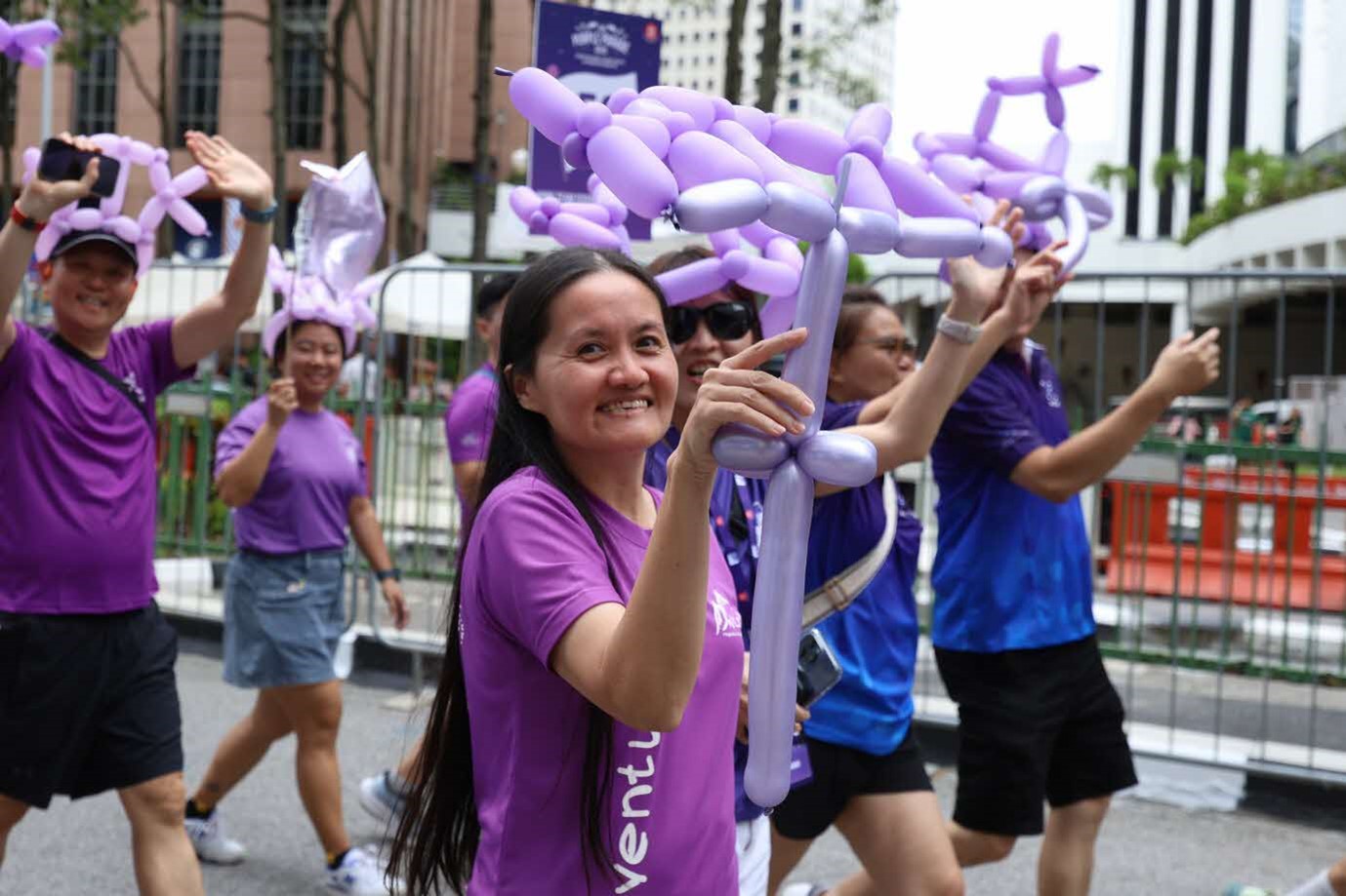 A lady in purple shirt raises a long purple balloon while walking amidst a crowd.