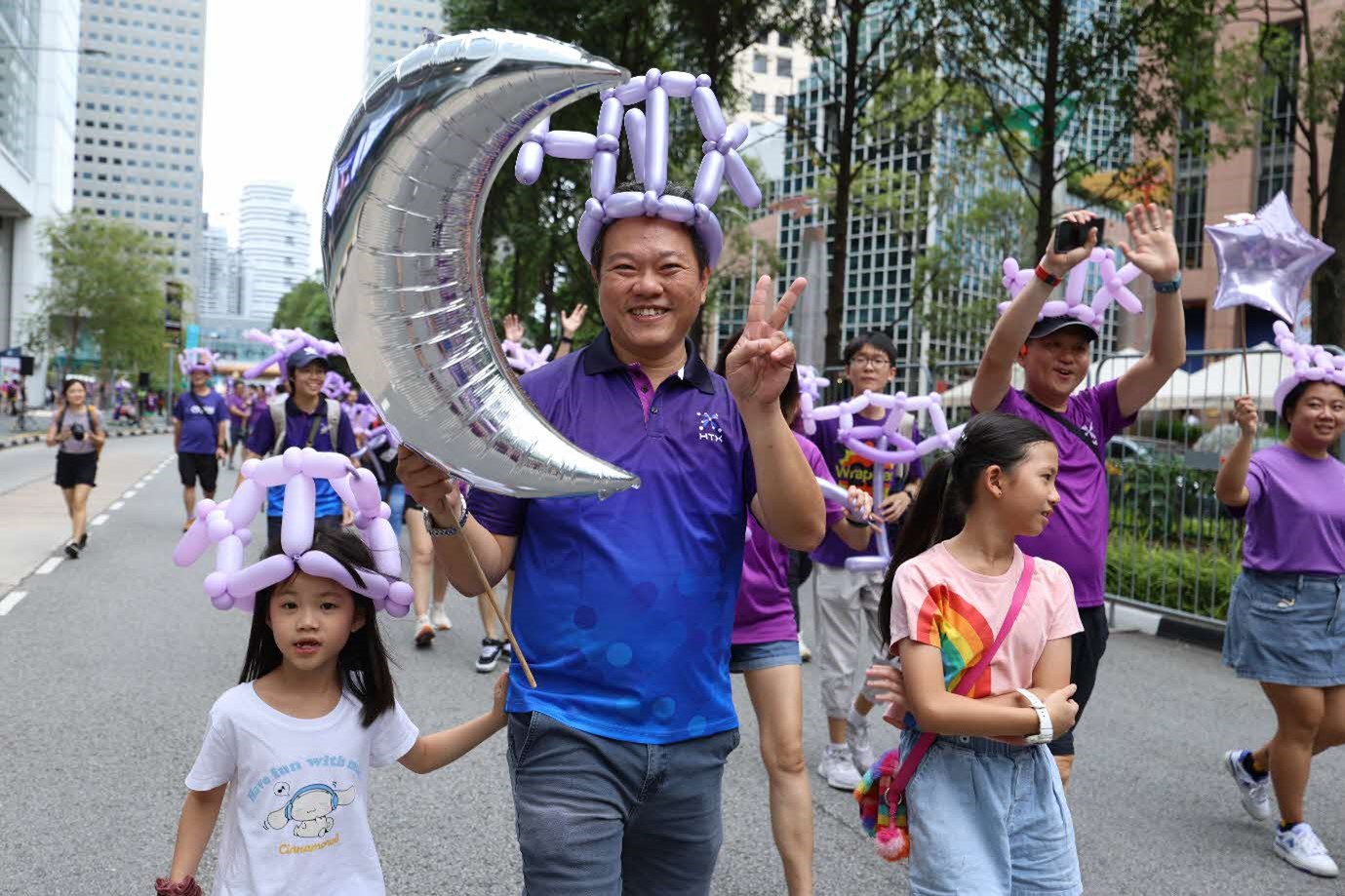 A man in a purple-and-blue polo shirt flashes a peace sign at the camera while carrying a silver crescent-shaped balloon and walking along two young girls.