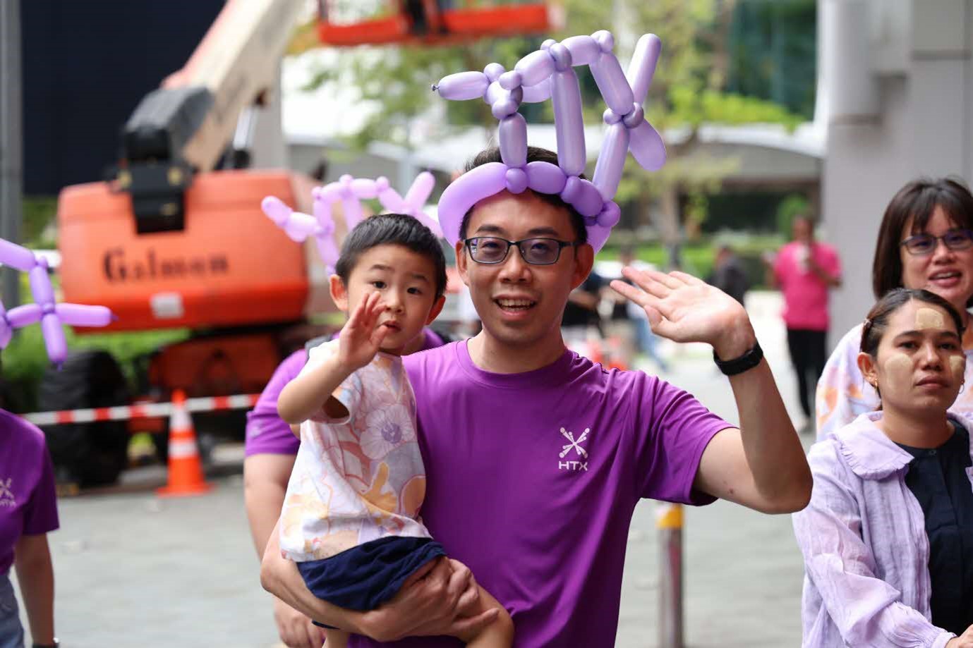 A male in purple shirt and balloon hat carries a young boy and they both wave to the camera.