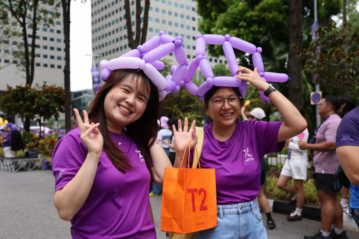 Two females in purple shirts and balloon hats wave to the camera.