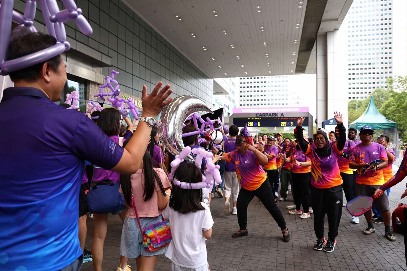 A man in a purple and blue shirt and wearing a balloon hat waves to a large group of people in purple and orange shirts, one of whom is waving back with both hands.