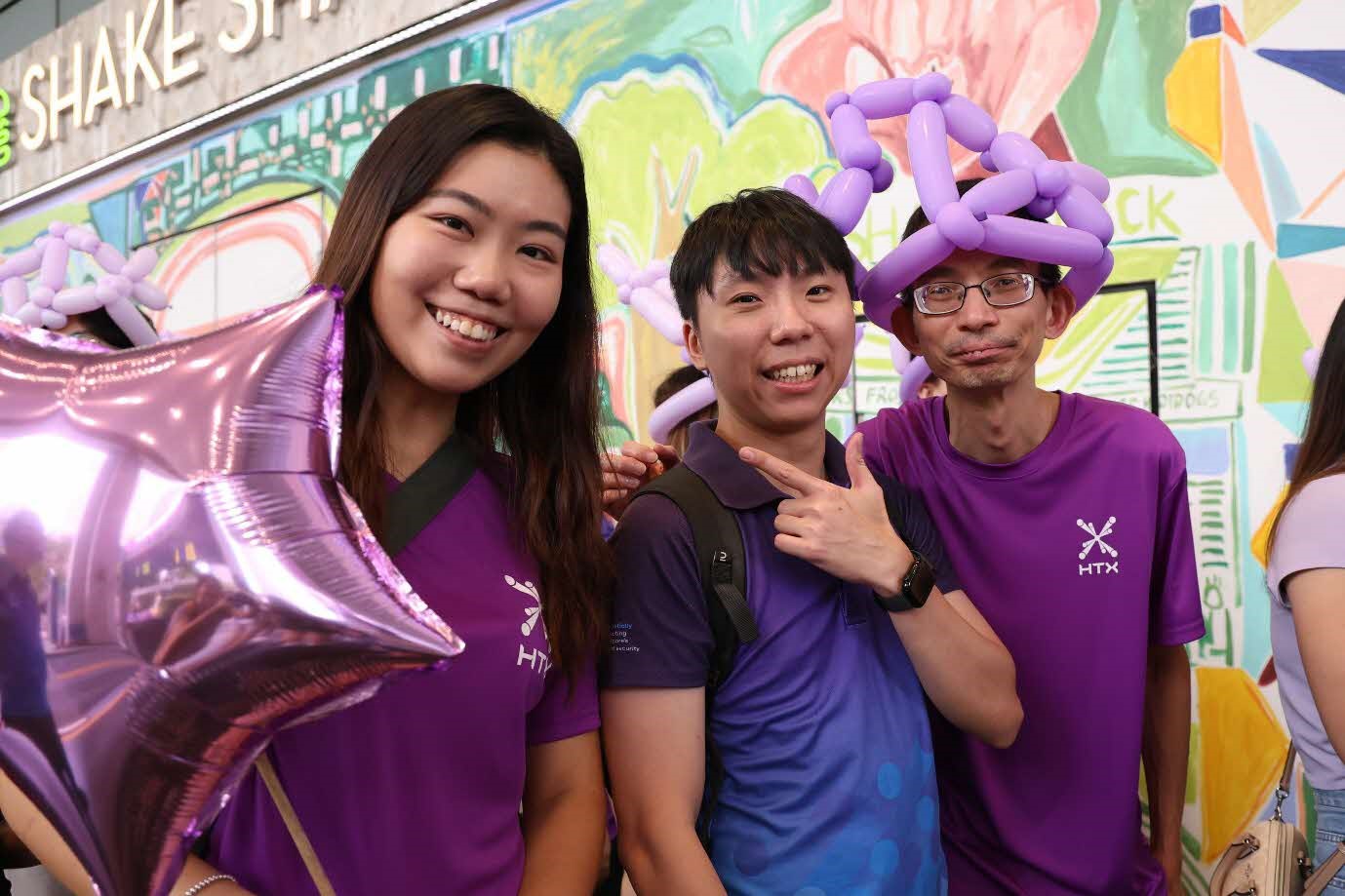 Three people in purple and blue shirts smile for the camera. One of them is holding a star-shaped balloon and another has a purple balloon hat.