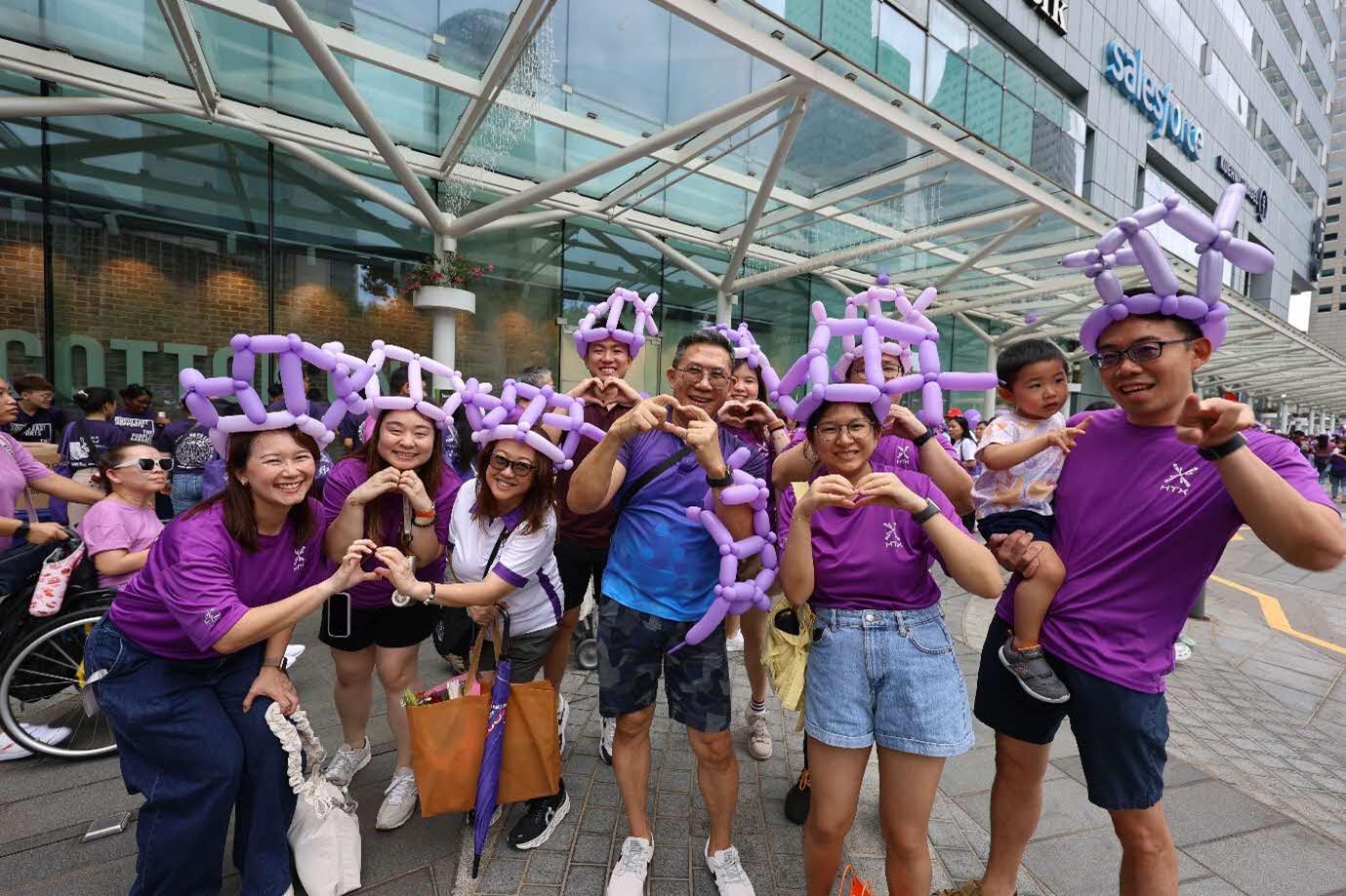 A group of people largely decked in purple shirts wearing purple balloon hats and making heart shapes with their hands.