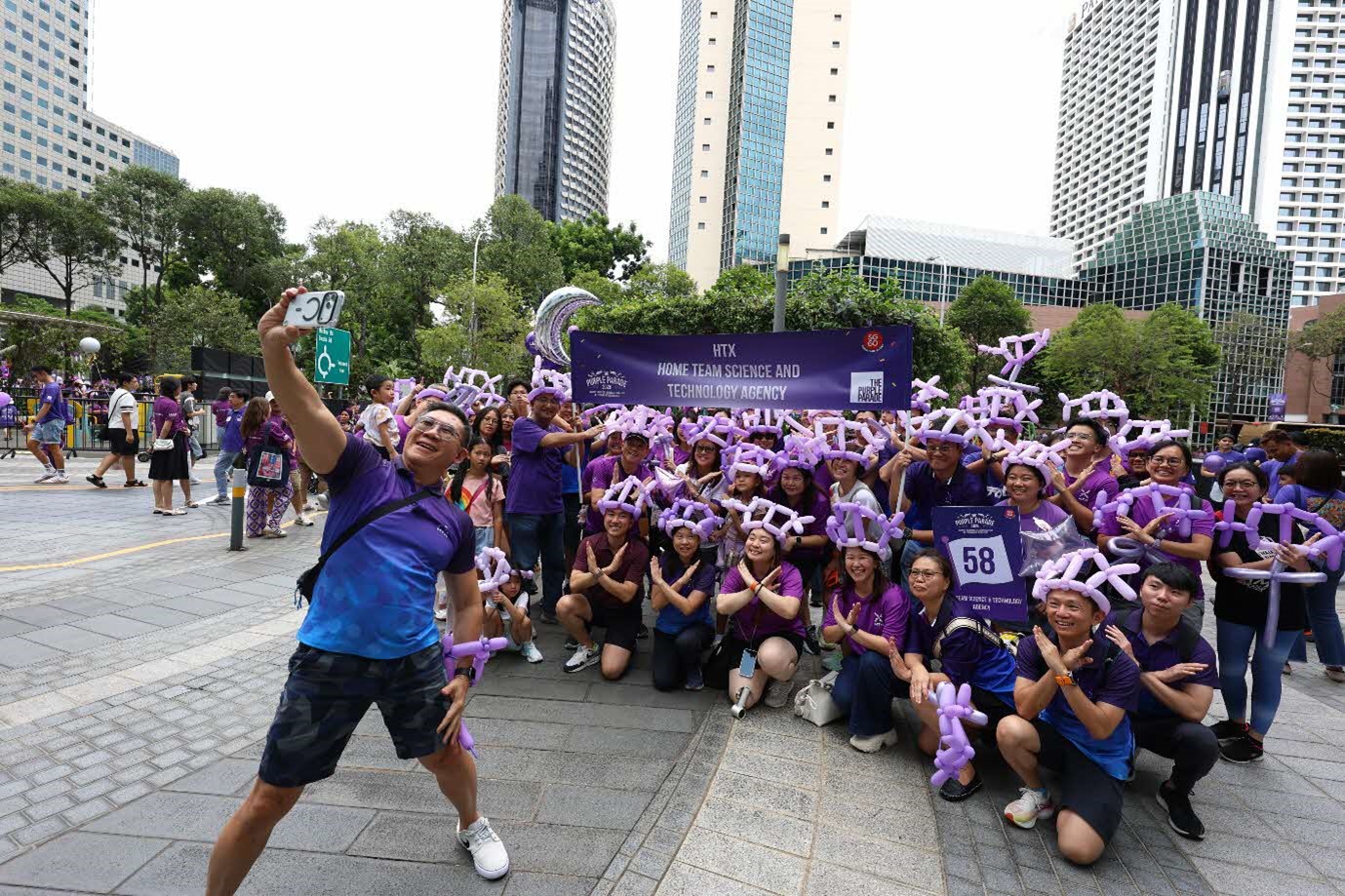A man holds a phone up for a selfie in front of a large group posing with their arms crossed and holding up a large banner that reads "HTX Home Team Science and Technology Agency".