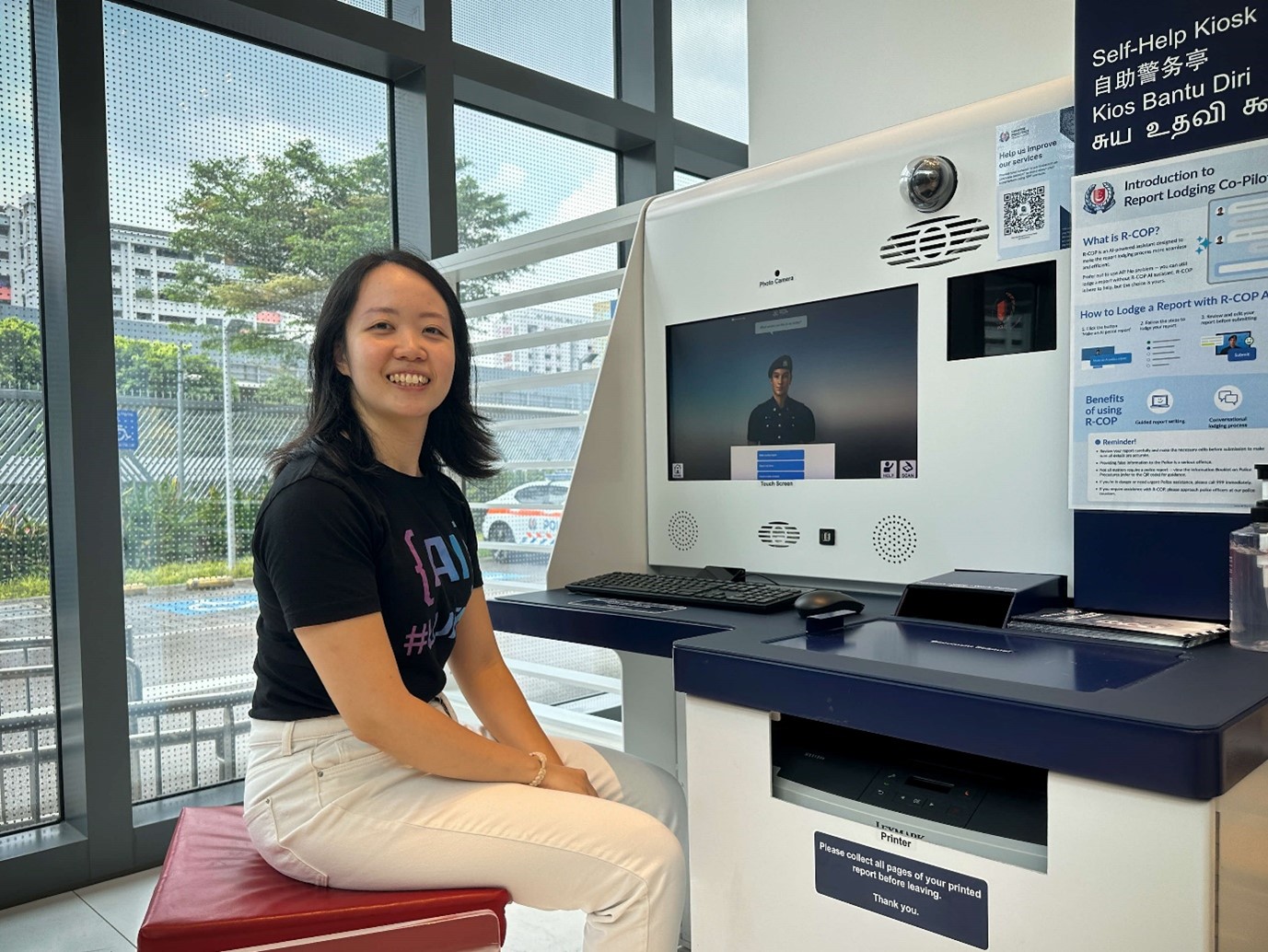 Woman sitting on a stool next to a police report kiosk, smiling at the camera.
