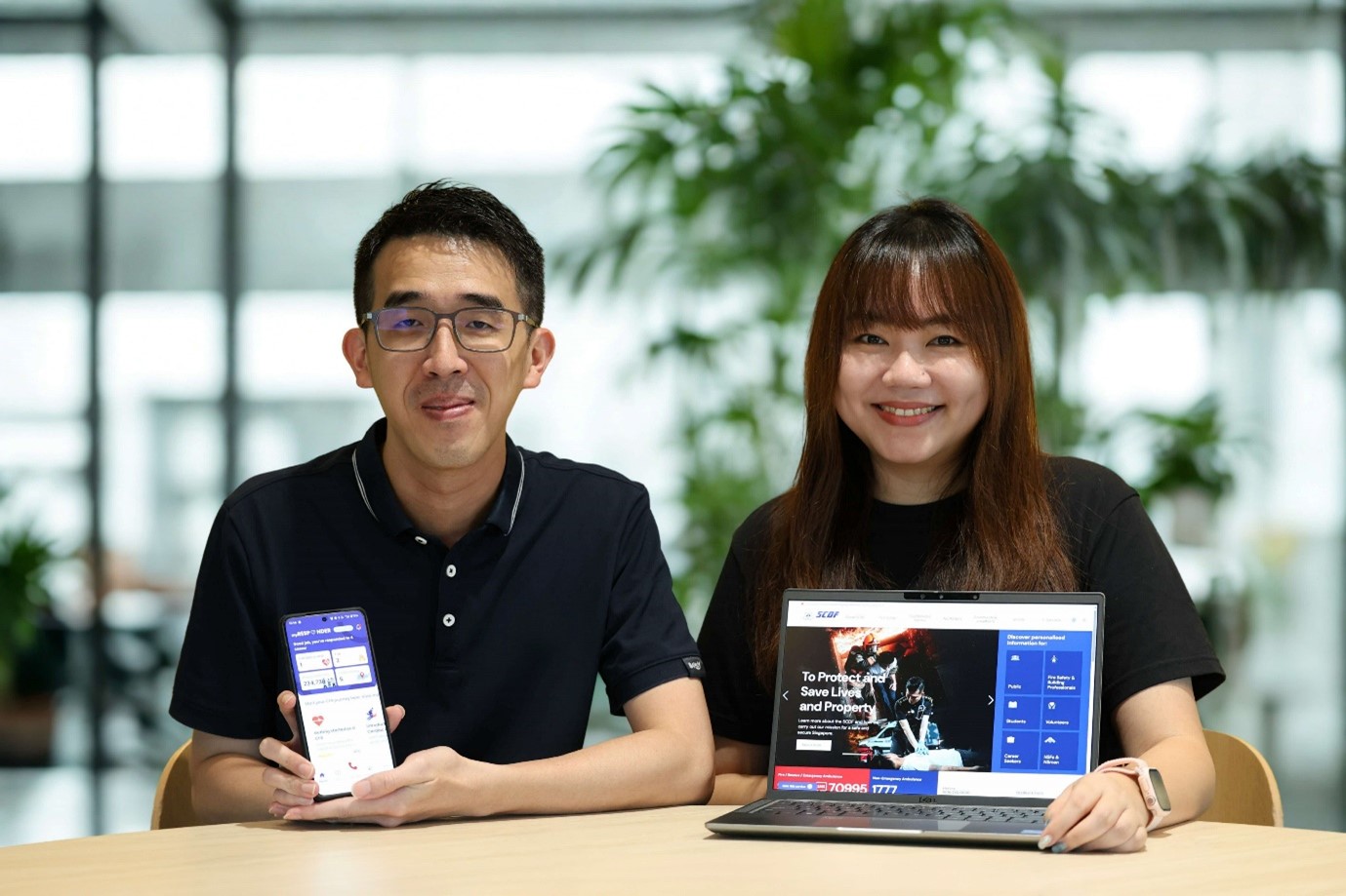 Two people sitting at a table, with a man holding a smartphone with the MyResponder app and a woman with a laptop displaying the SCDF website.