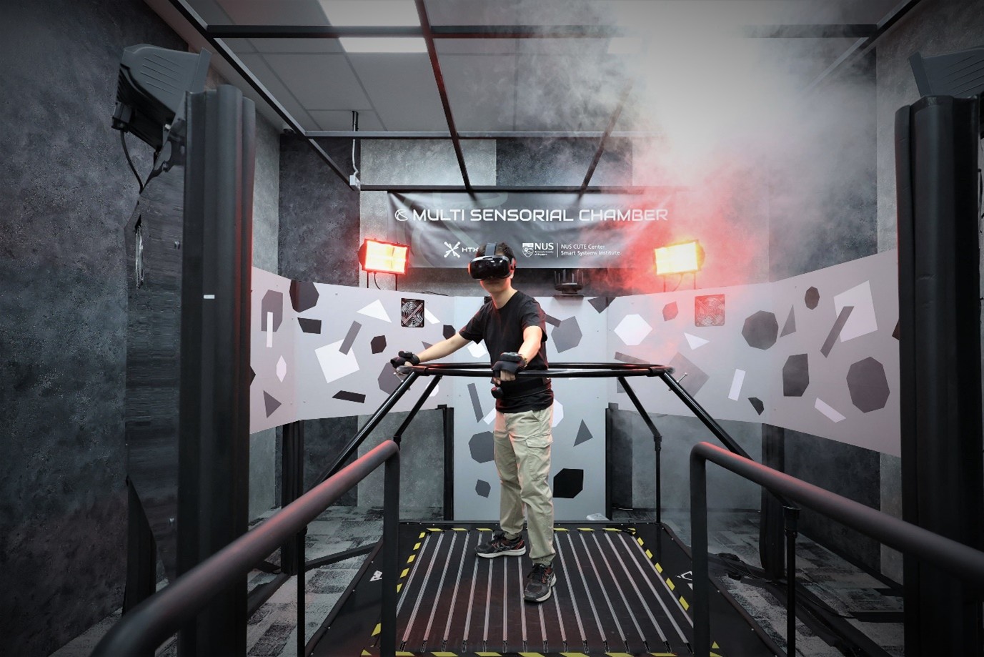 A person wearing virtual reality goggles, standing on a treadmill with railings all around and smoke and red lights in the environment.