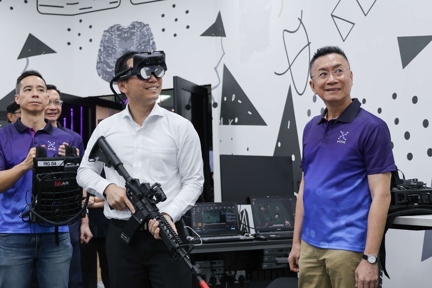 MOS Goh in a white shirt and VR goggles holds a rifle while standing next to another man in a purple HTX shirt, with other people and technical equipment in the background.