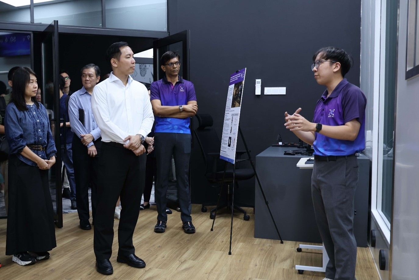 A group of people, including MOS Goh, listen as an officer in HTX's purple shirt gestures and speaks next to a presentation poster in a room.