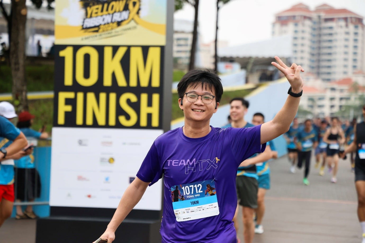 Smiling male runner in purple Team HTX shirt raising a victory hand sign near a "10km Finish" sign.