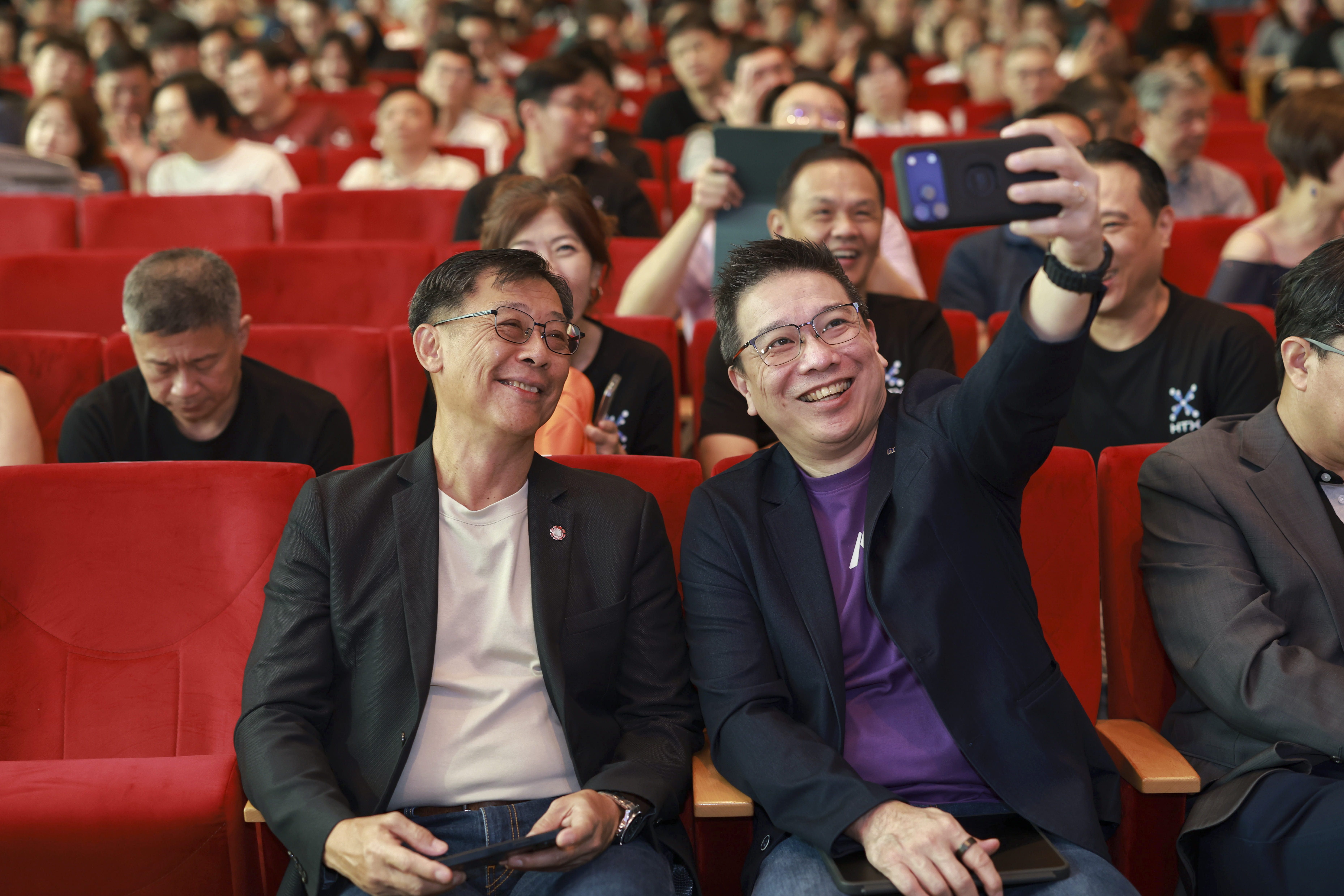 Two smiling men seated in red auditorium chairs taking a selfie.