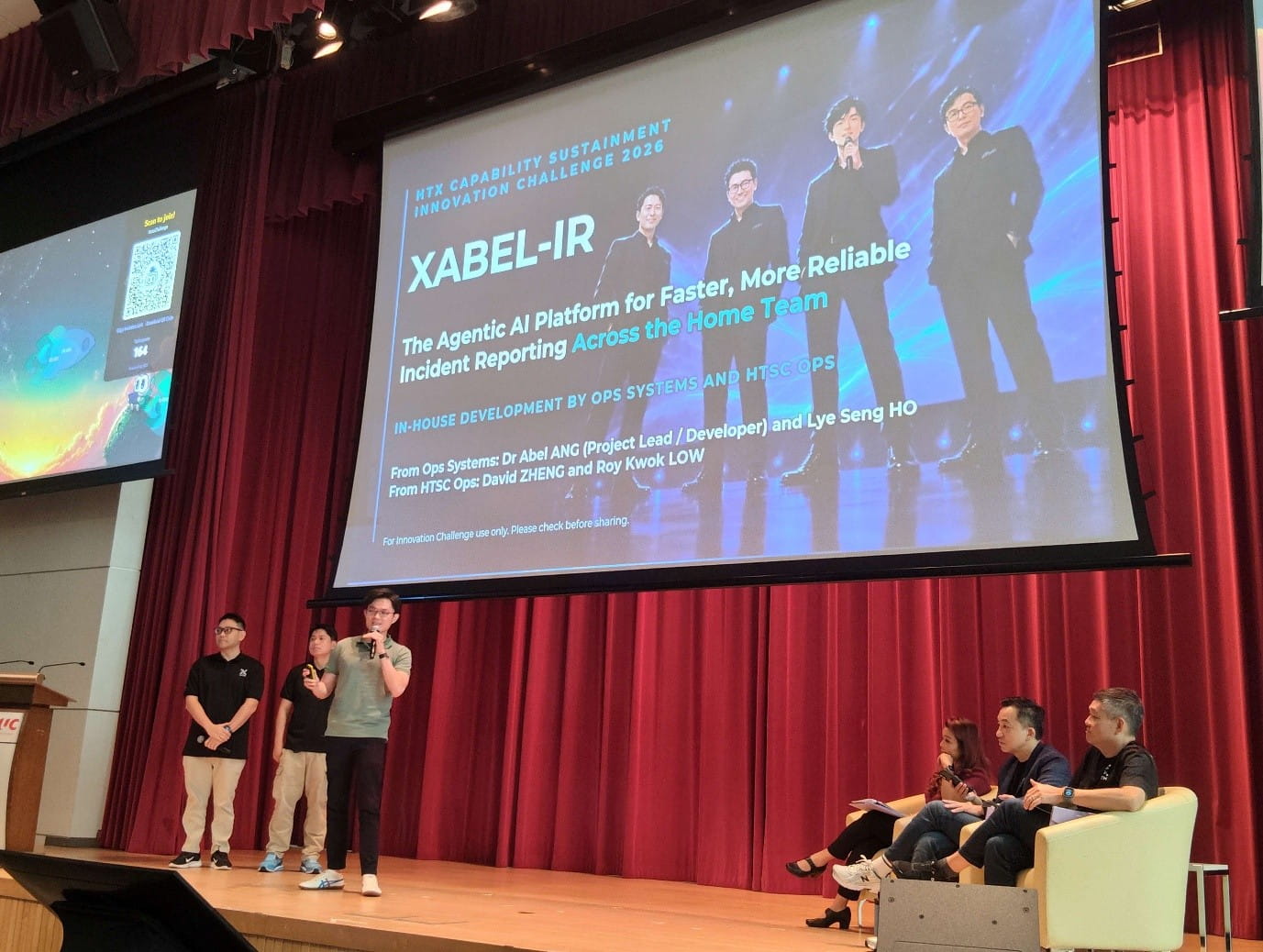 Stage photo showing a team presentation for, while presenters stand onstage with seated panellists.