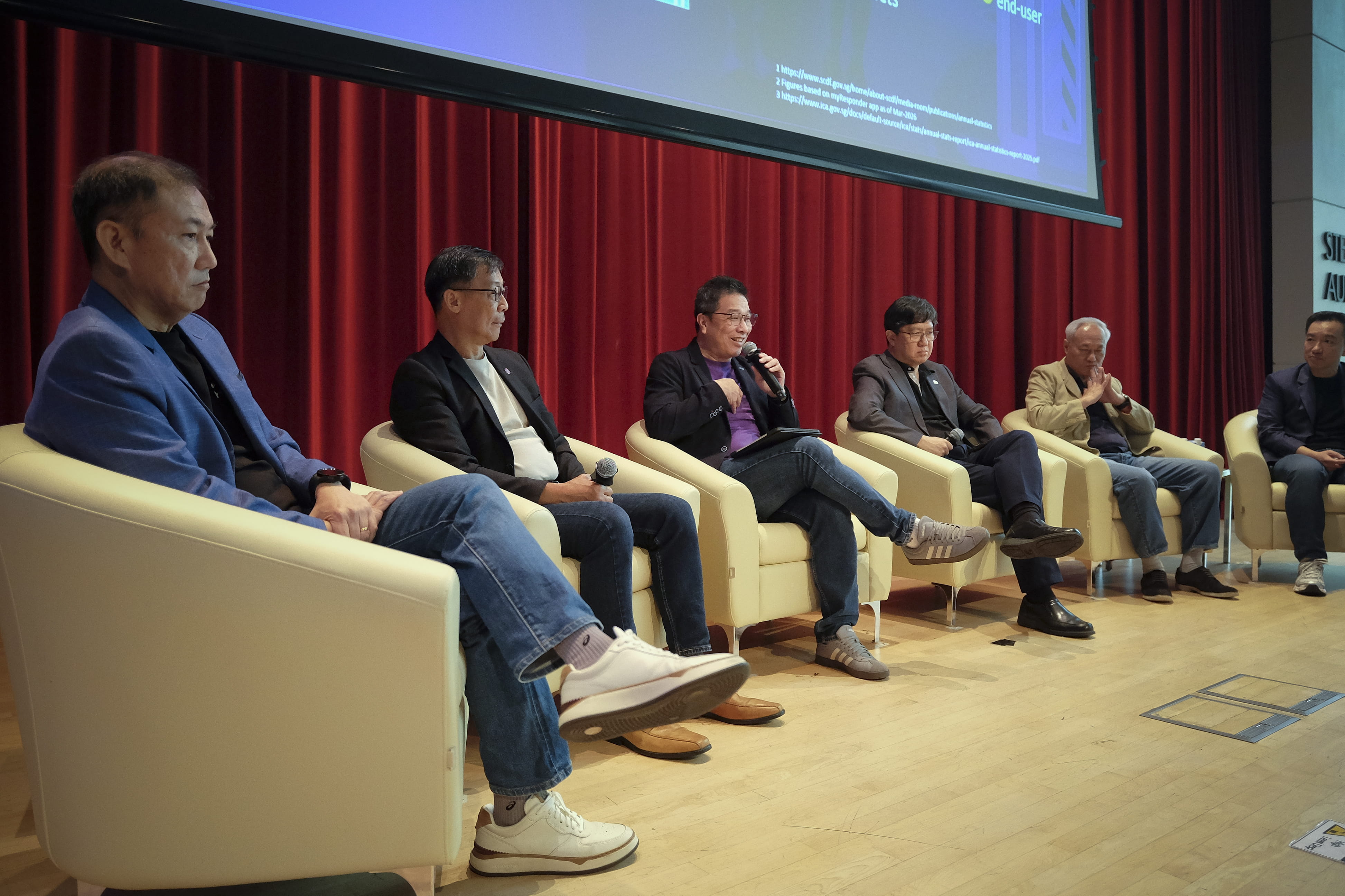Six panellists seated in armchairs on stage during a discussion.