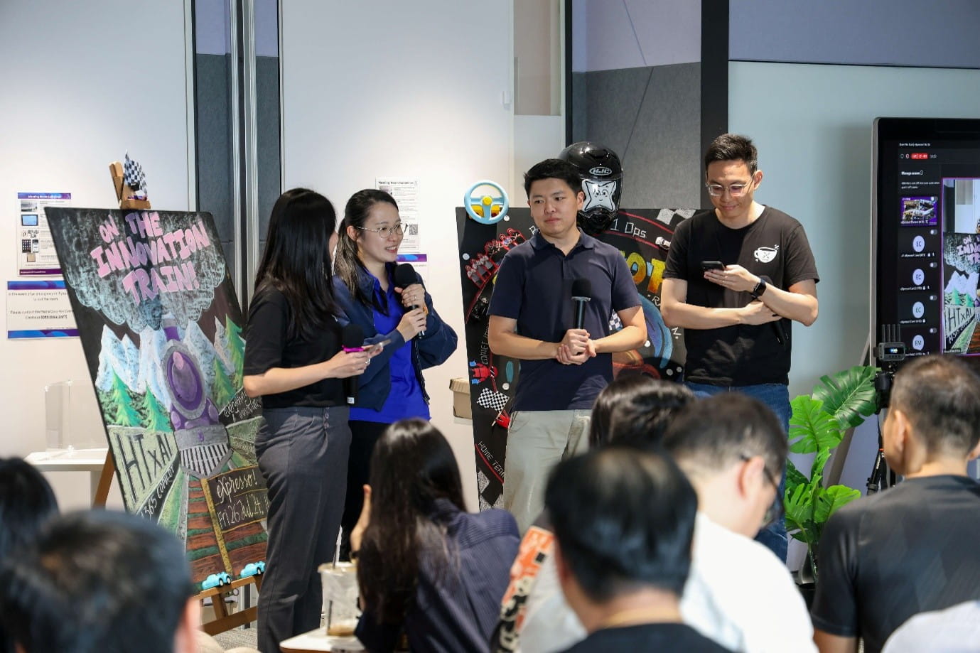 Two presenters addressing a seated audience indoors, with two emcees by their side.