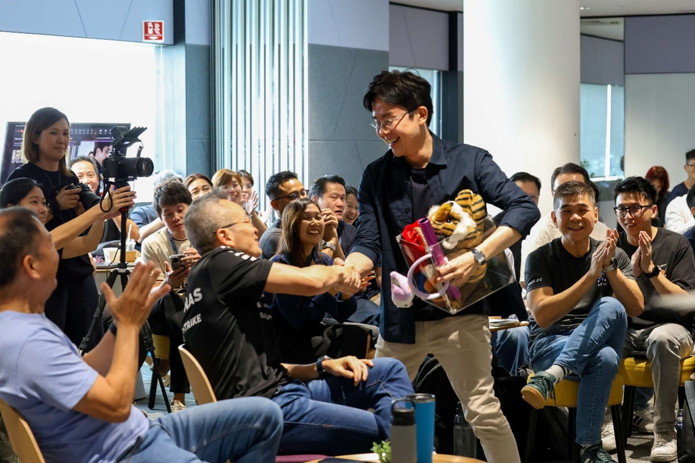 A smiling man carrying a gift bag while shaking another man's hand among the live audience.