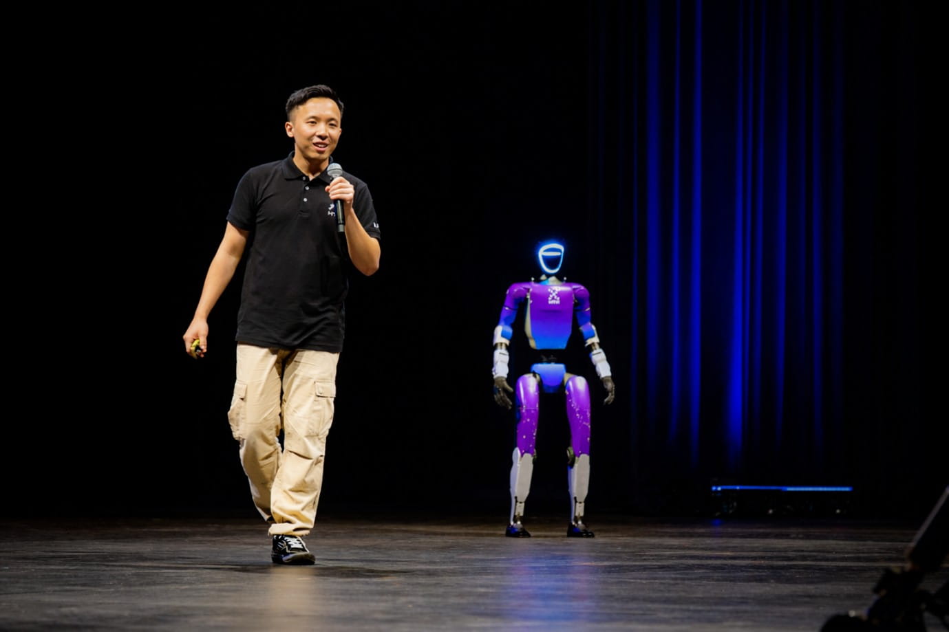 Convention 11 A man walking on stage next to a humanoid robot with purple and white body and illuminated blue face.