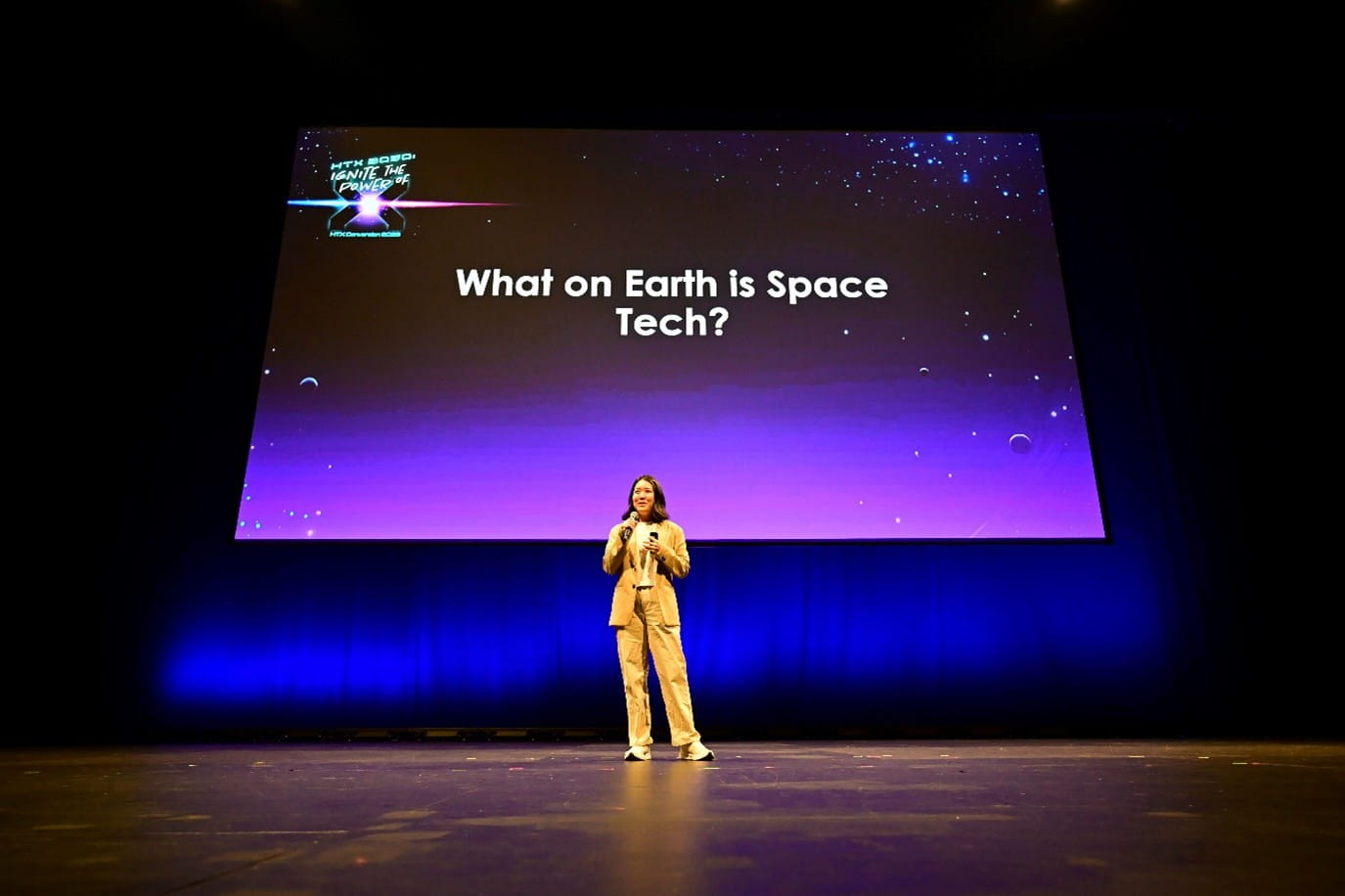 A speaker standing on stage in front of a large screen displaying the text "What on Earth is Space Tech?"