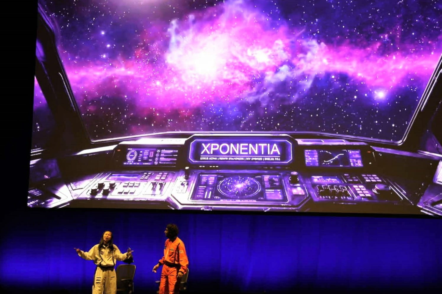 Two presenters standing on stage in front of a large screen displaying a spaceship cockpit with a vibrant purple galaxy outside. The cockpit dashboard features the word "XPONENTIA" prominently.