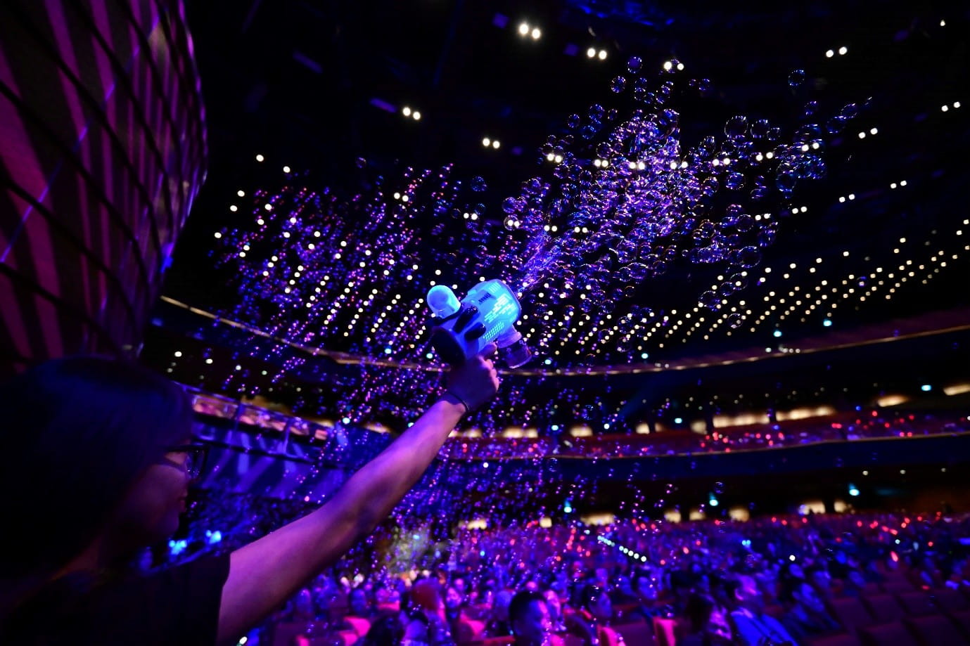 A person spraying a bubble gun towards a large crowd illuminated by purple and blue stage lights.