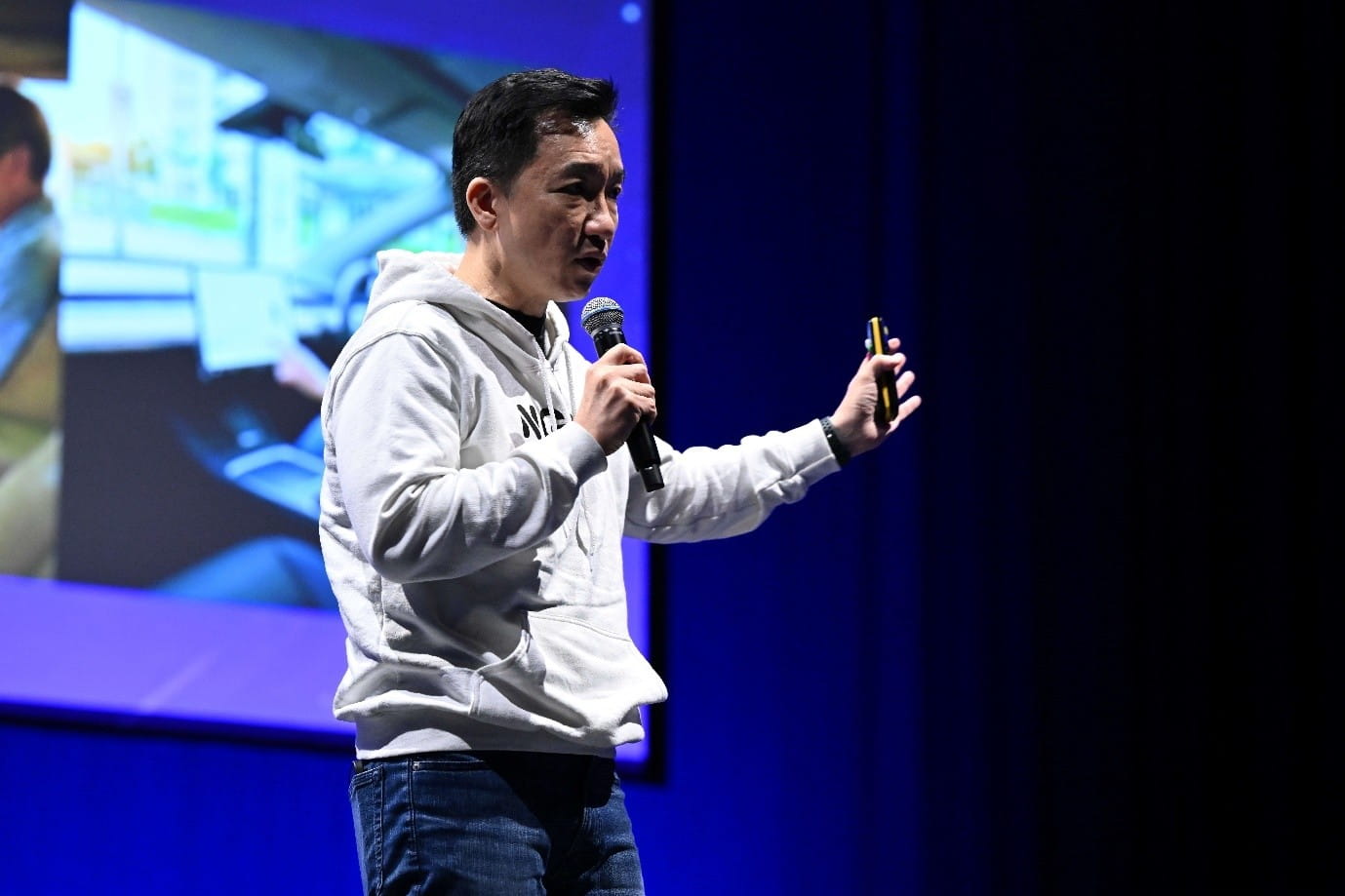 A man in a white hoodie and jeans speaks on stage with a microphone in one hand and a remote control in the other.