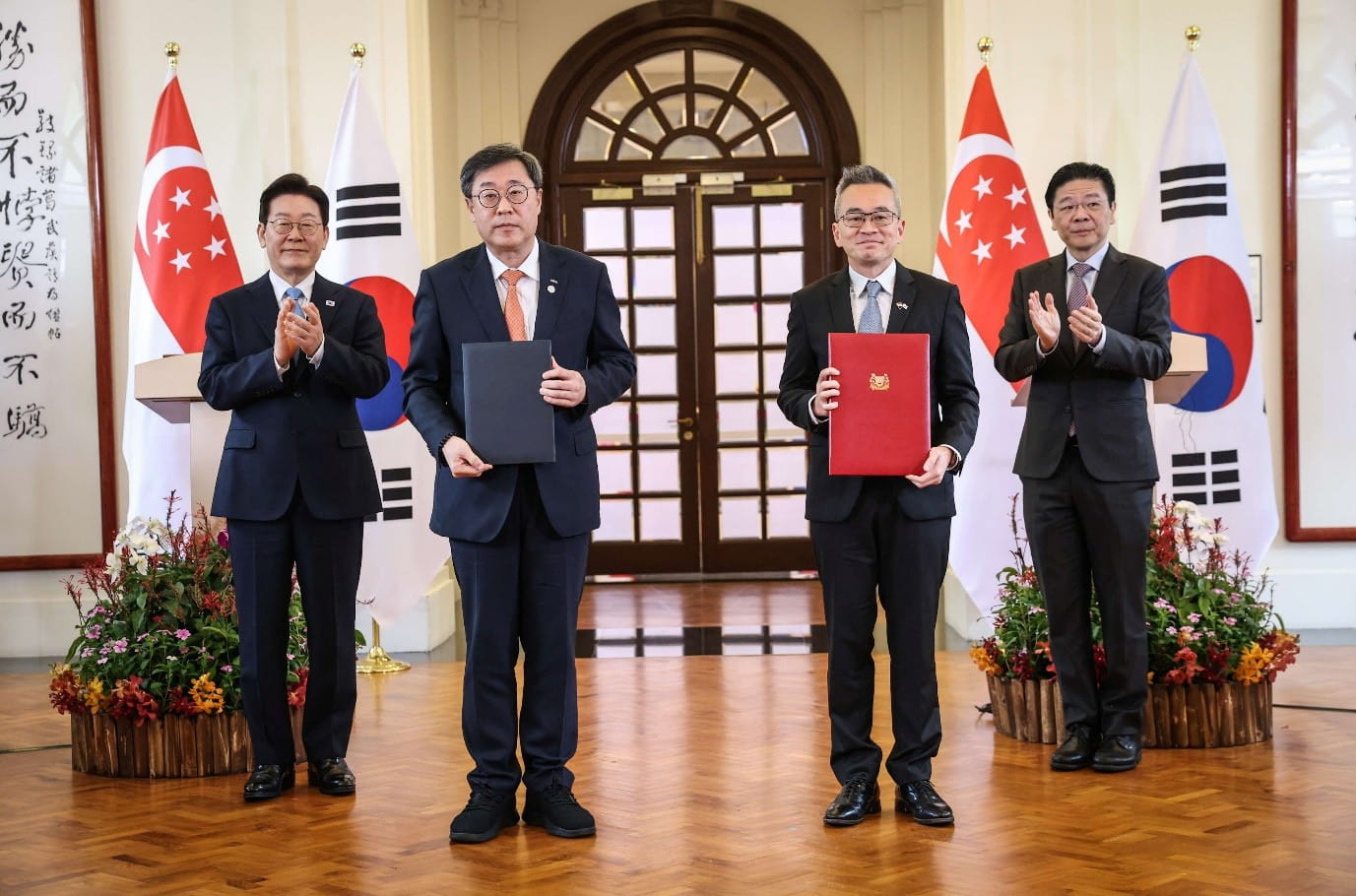 Four officials pose with the flags of Singapore and South Korea. Two in the centre hold signed folders, marking a formal agreement.