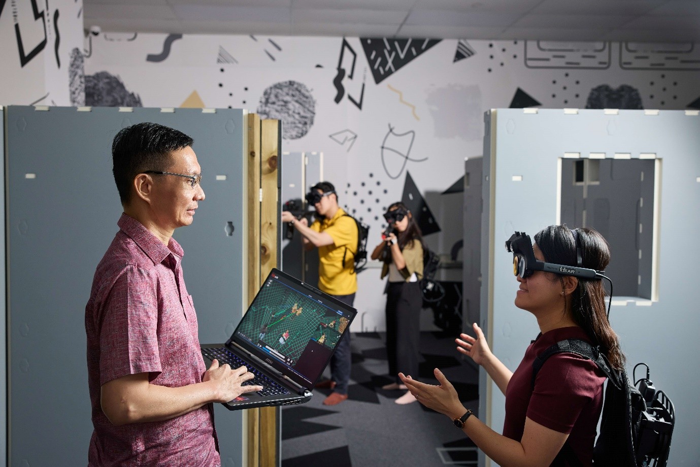 Two people in the foreground discuss something while one carries a laptop with a 3D map of the room and one is wearing an equipment backpack and goggles. In the background, two people are wearing the same equipment and carrying mock rifles.
