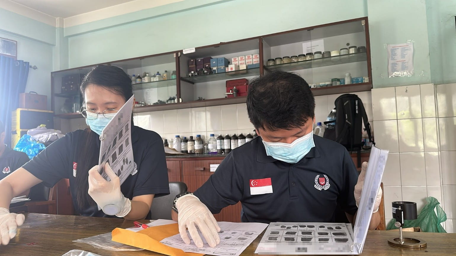 Two masked officers reviewing paperwork and samples at a workbench.
