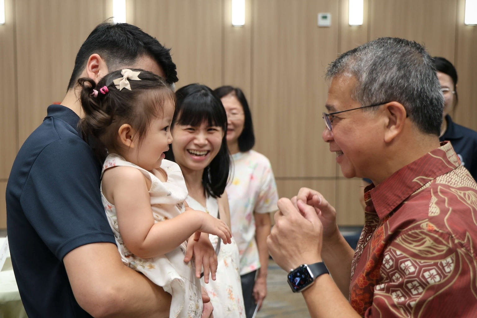 A man holding a young girl while an older man smiles and gestures toward her.
